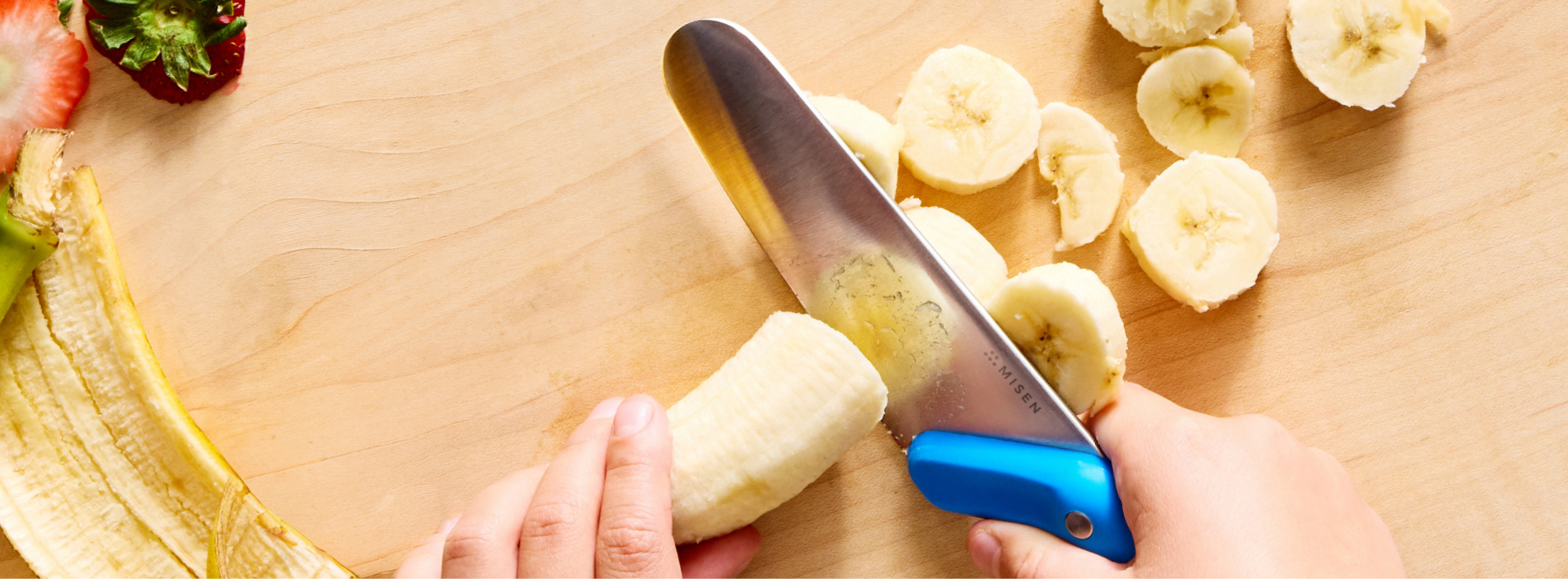An overhead shot shows a pair of hands using a blue-handled Misen Kids' Knife to slice a peeled banana on a light wooden cutting board. Several slices of banana are scattered to the right of the knife, and the banana peel is visible on the left. A sliced strawberry is partially visible in the upper left corner. The word "MISEN" is visible on the knife blade near the handle.