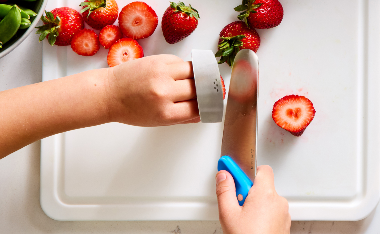 An overhead shot showing a child's hands slicing strawberries on a white cutting board. The right hand holds a Misen Kids' Knife with a blue handle and a stainless steel blade. The left hand wears a stainless steel finger guard while holding a strawberry steady. Several whole and sliced strawberries are visible on the cutting board and near the upper left. The word "MISEN" is visible on the knife blade.