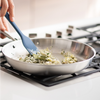Stainless steel skillet on gas burner with chopped onions, herbs, and a bay leaf being stirred with a blue silicone spatula.