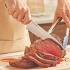 Two hands using a Misen knife and tongs to slice medium-rare roast on wooden cutting board