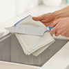 Two hands drying a wet chef's knife with water droplets using a folded white cloth over a kitchen sink.